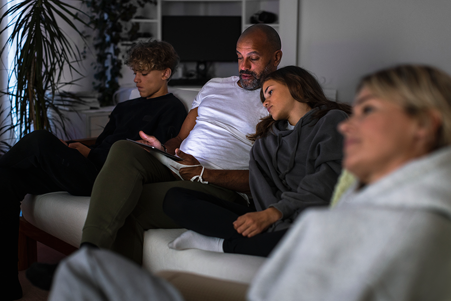 En familj sitter i soffan. Mannen sitter med två barn och tittar på en surfplatta. Kvinnan tittar på TV.